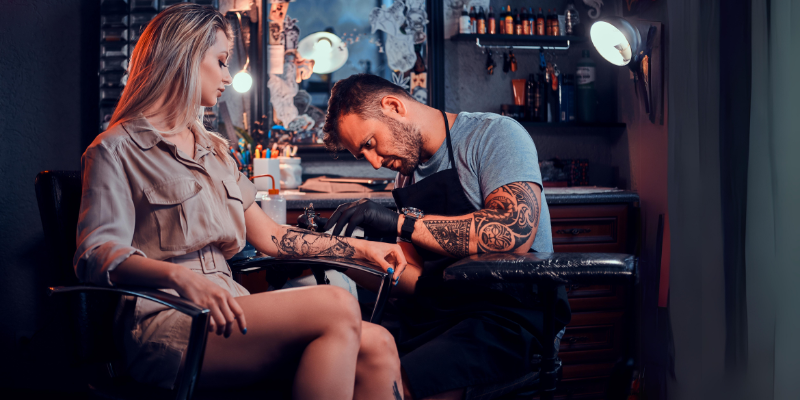 Eine Frau bekommt ein Tattoo von einem Tattoo-Artist, man sieht den Arbeitsplatz des Tätowierers und den Vorgang des Tätowierens.